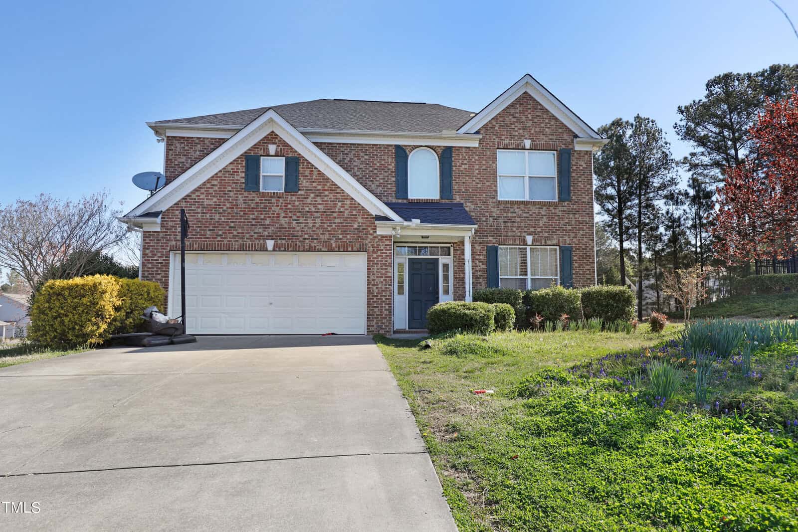 Located at 2108 Westbourne Drive in Creedmoor, this two-story brick house with white trim boasts a two-car garage and a spacious driveway. A lush grassy front yard, dotted with bushes and set against a backdrop of trees, completes the charming scene under clear, sunny skies.