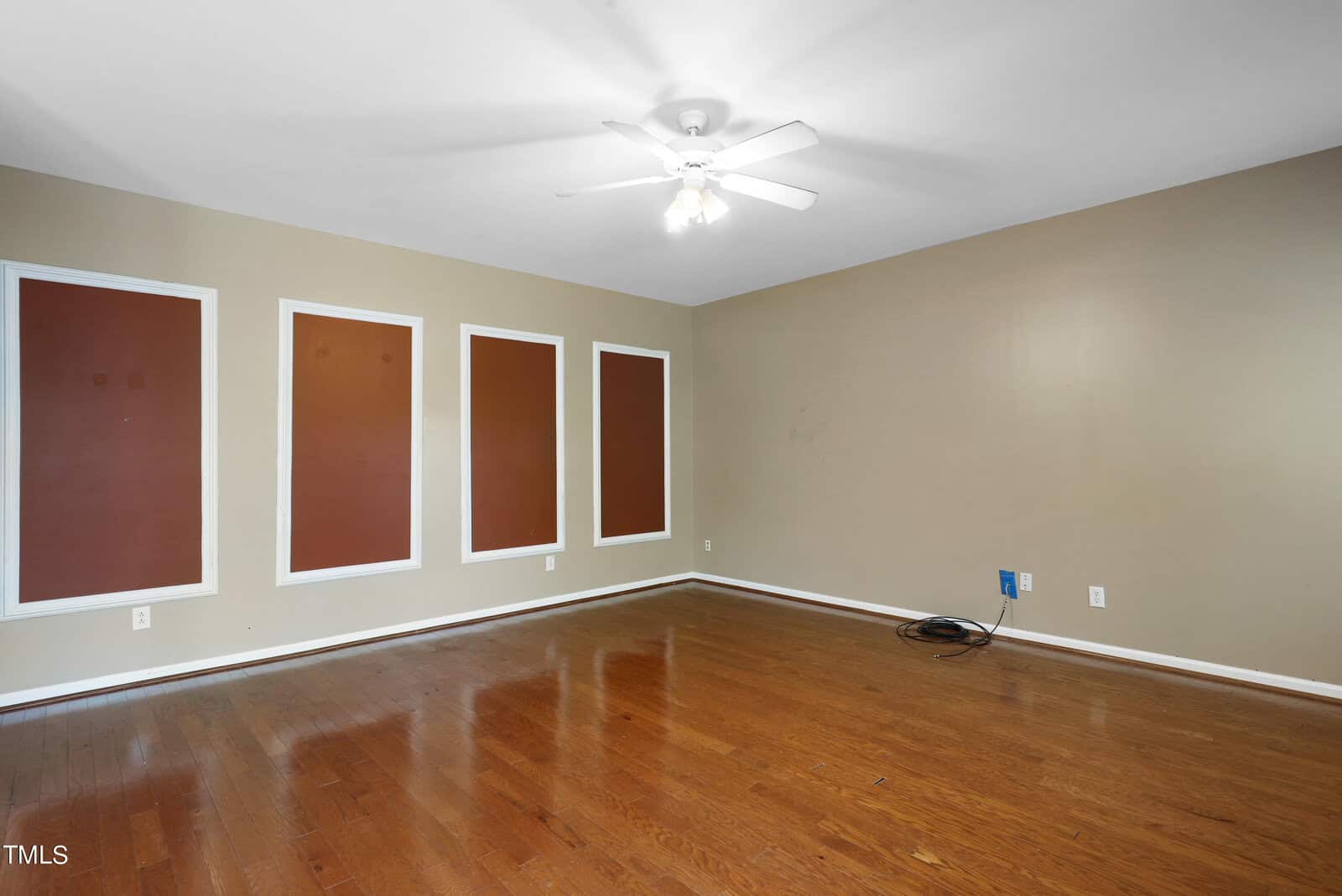 An empty room with wooden floors and beige walls features four brown rectangular panels framed in white. A ceiling fan is centered above, and there is a bundle of cables near one wall outlet.