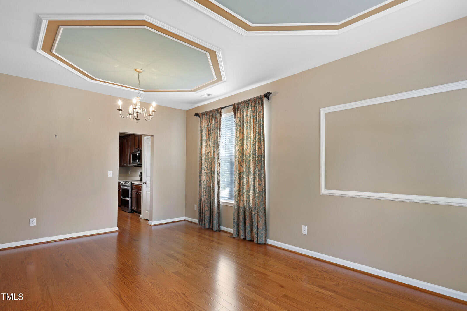 A spacious, empty dining room with hardwood floors, beige walls, and a decorative ceiling with two octagonal accents. A light fixture hangs above, and a window features floral curtains. An open doorway leads to a visible kitchen area.
