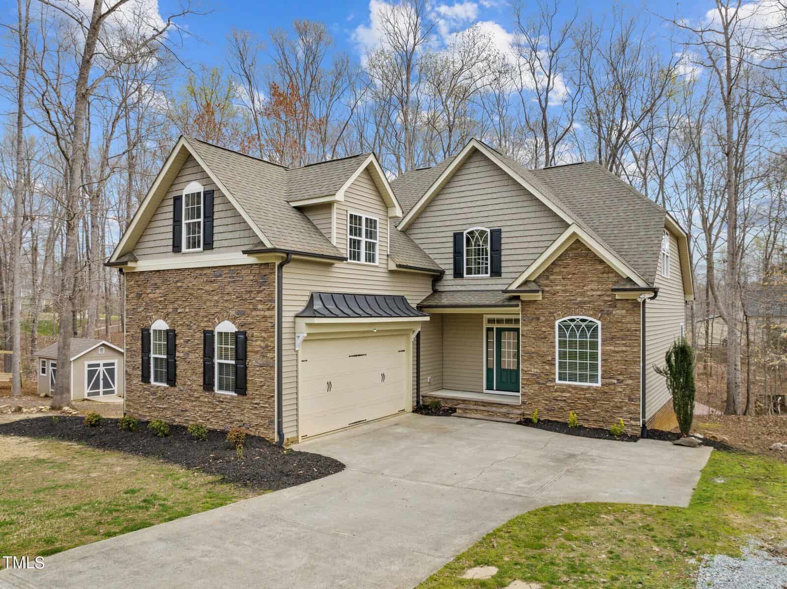 A two-story house with a mix of stone and beige siding, featuring a two-car garage and an arched window. The home is set against a backdrop of leafless trees and a small wooden shed is visible to the left. A concrete driveway leads to the entrance.