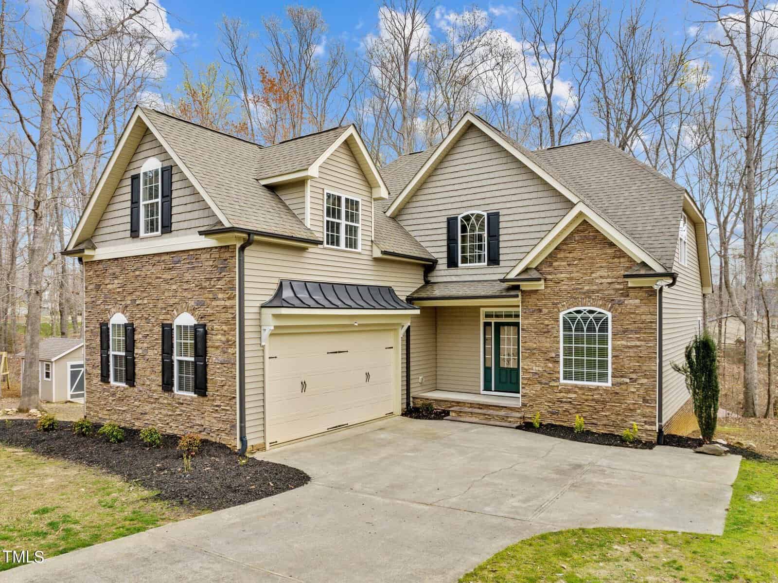 A two-story suburban house with beige siding and stone accents. It has a double garage, black shutters, and a front porch. The yard is partially landscaped, and the background features leafless trees and a clear blue sky.
