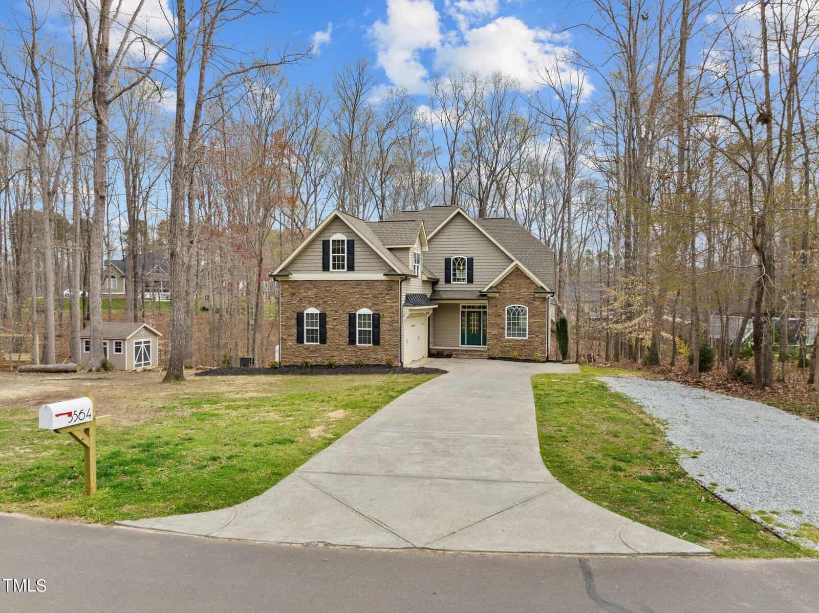 A two-story brick house with a double garage and a wide driveway. The yard is bordered by tall, leafless trees, and a white mailbox labeled "364" stands near the road. The sky is clear with a few clouds.