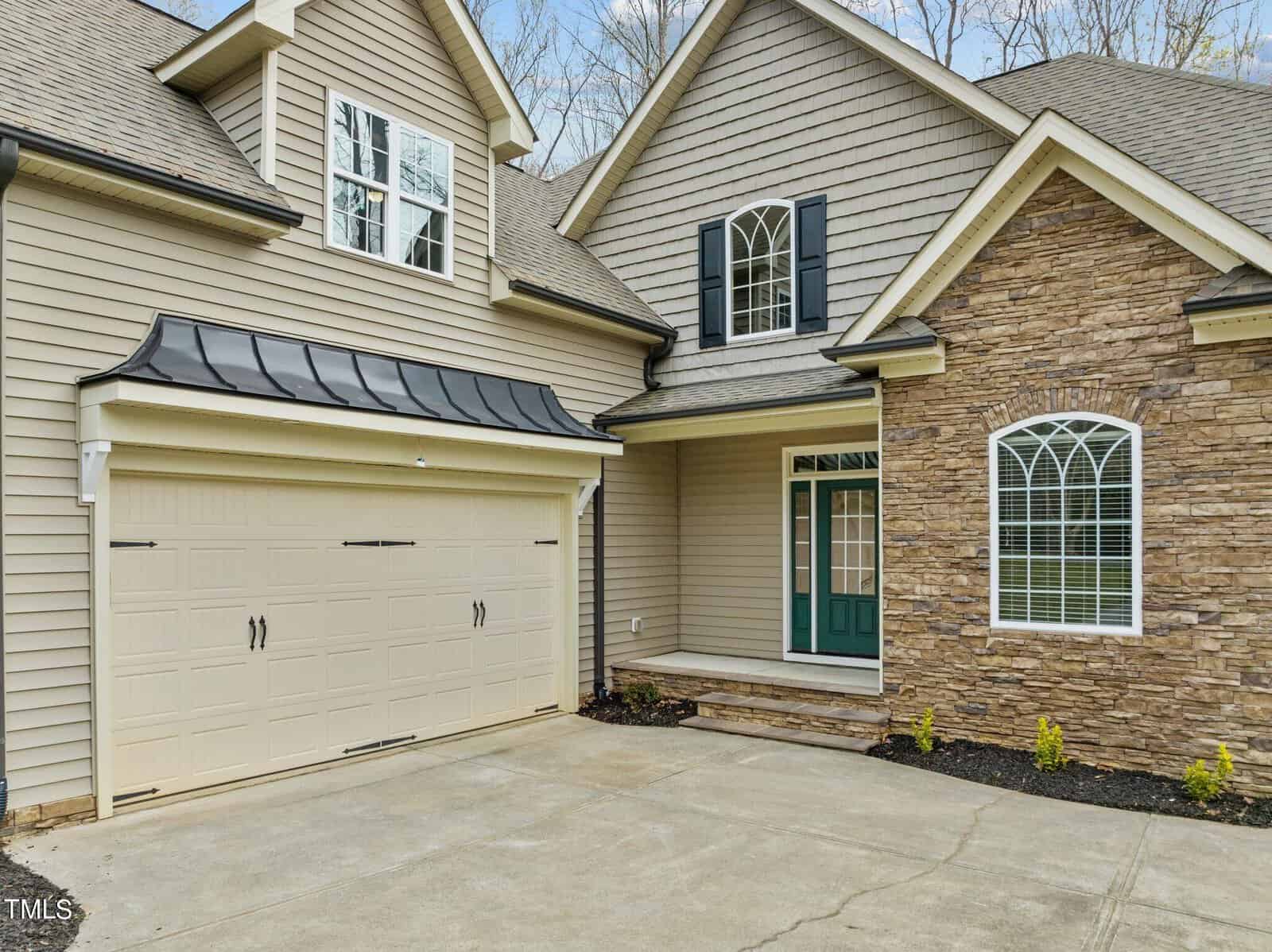 A modern suburban house with beige siding and stone accents. The house features a two-car garage with decorative doors, arched windows, and a small covered entrance. The driveway is concrete, and there are minimal landscaping details.