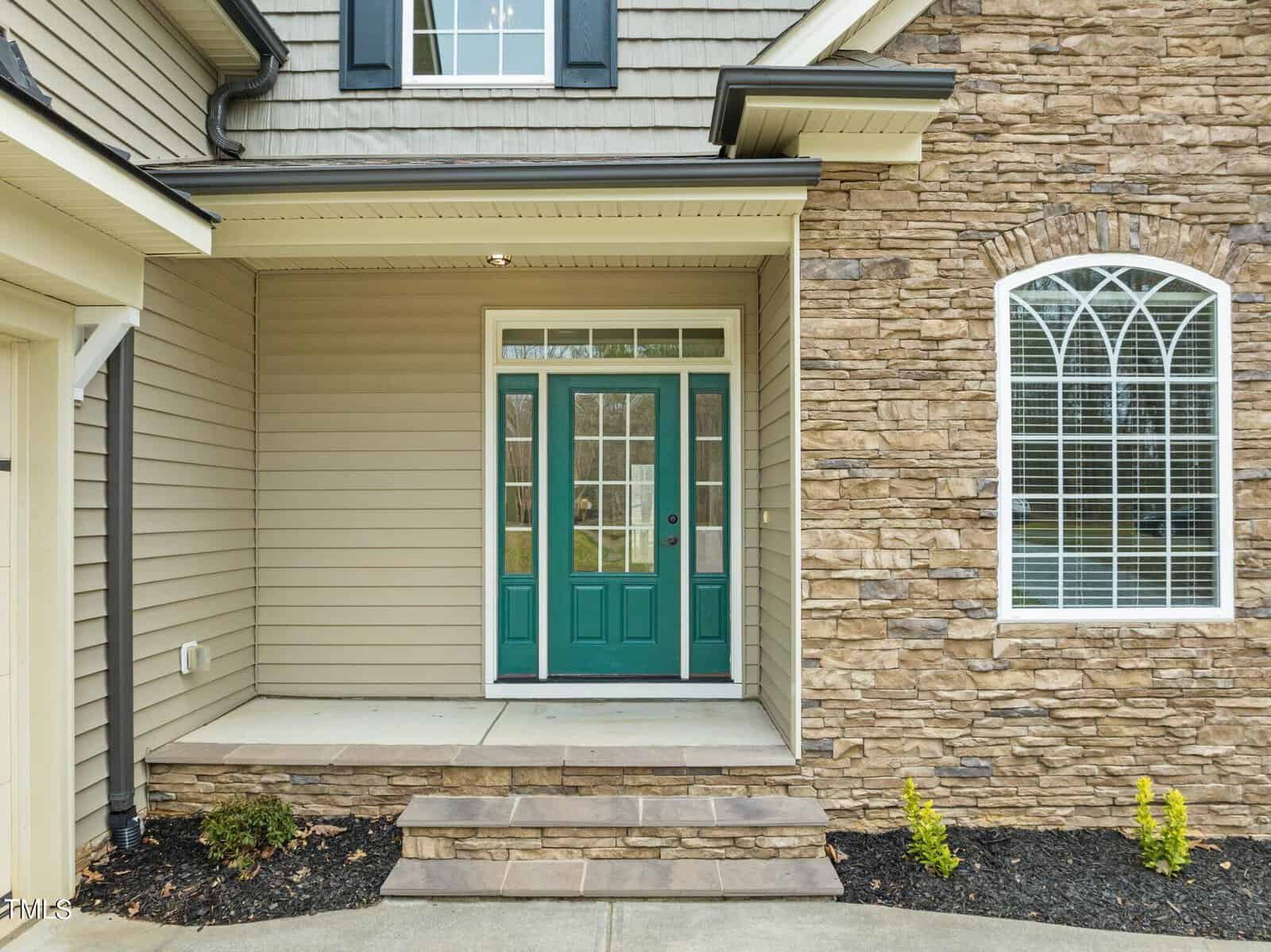 Front porch of a house with stone and siding exterior, featuring a green door with glass panels and two sidelights. An arched window is on the right above stonework, and the entrance is accessed via a small step with a concrete path.