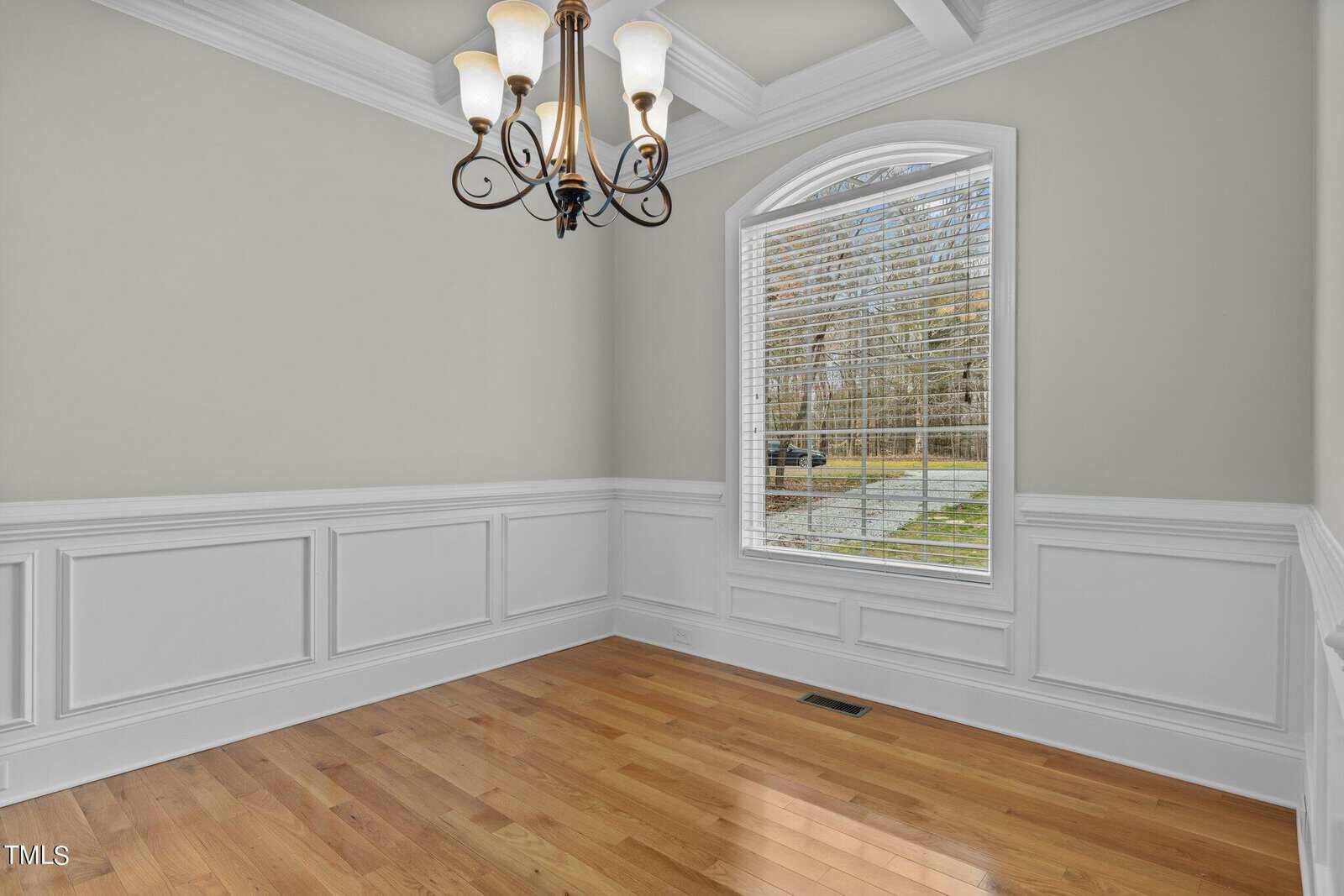 A bright, empty room with light green walls and white wainscoting. There's a large arched window with blinds, a hardwood floor, and a ceiling-mounted chandelier with multiple lights.