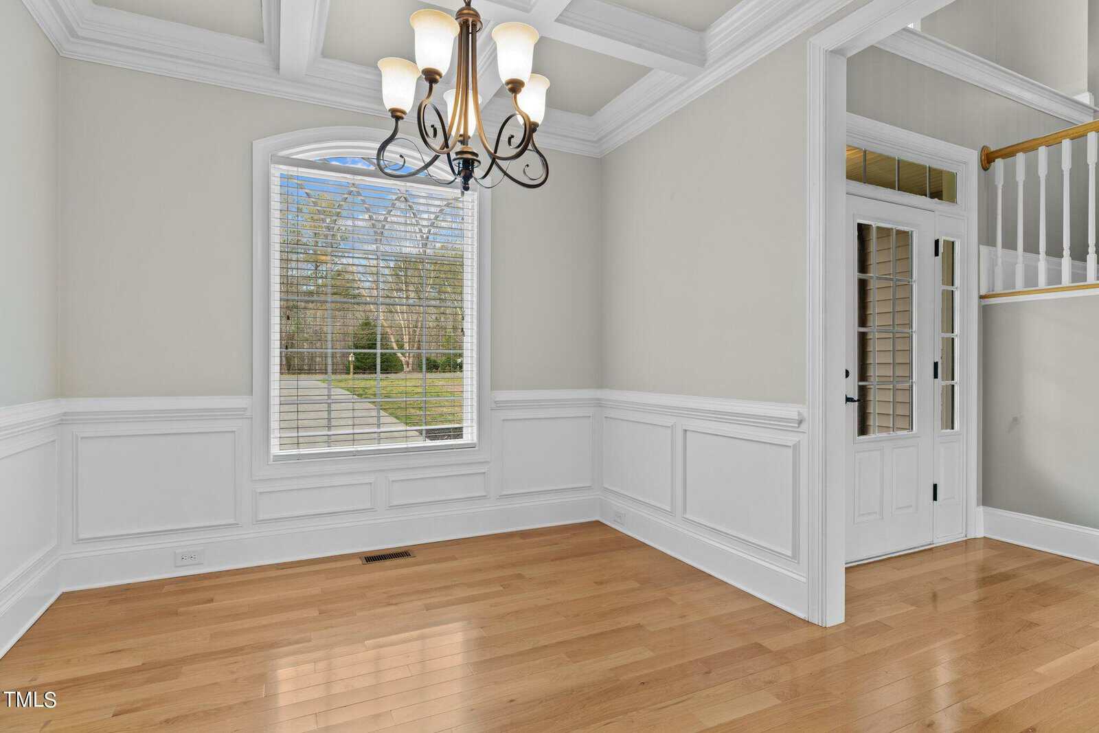 Elegant empty room with hardwood floors, light green walls, and white wainscoting. A large window with blinds overlooks a garden. A black chandelier hangs from a coffered ceiling, and a white door with glass panels is on the right.
