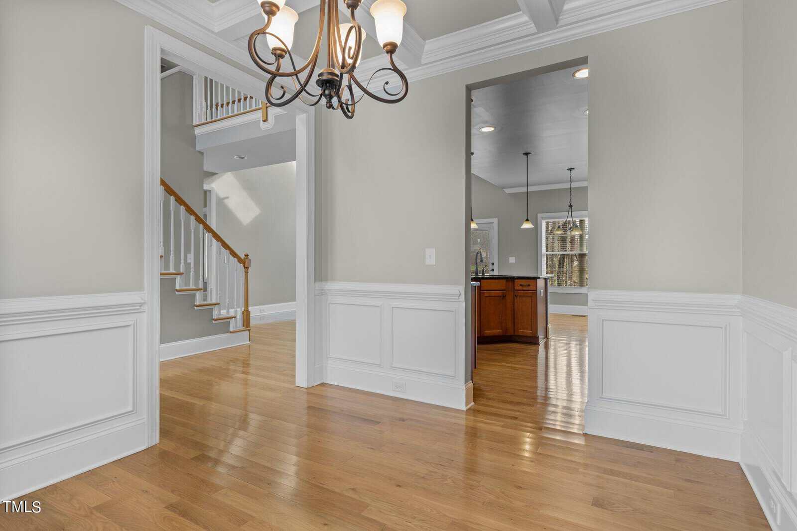 A bright, spacious interior features a hallway with light green walls and white wainscoting, leading to a kitchen with wood flooring. The space includes an elegant chandelier and a wooden staircase in the background. Natural light streams in through windows.