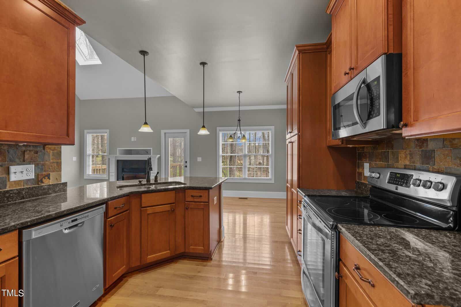 Kitchen with wooden cabinets, black granite countertops, stainless steel appliances, and a slate backsplash. The space features hardwood floors and pendant lighting, with large windows providing natural light. An opening leads to a living area with a fireplace.