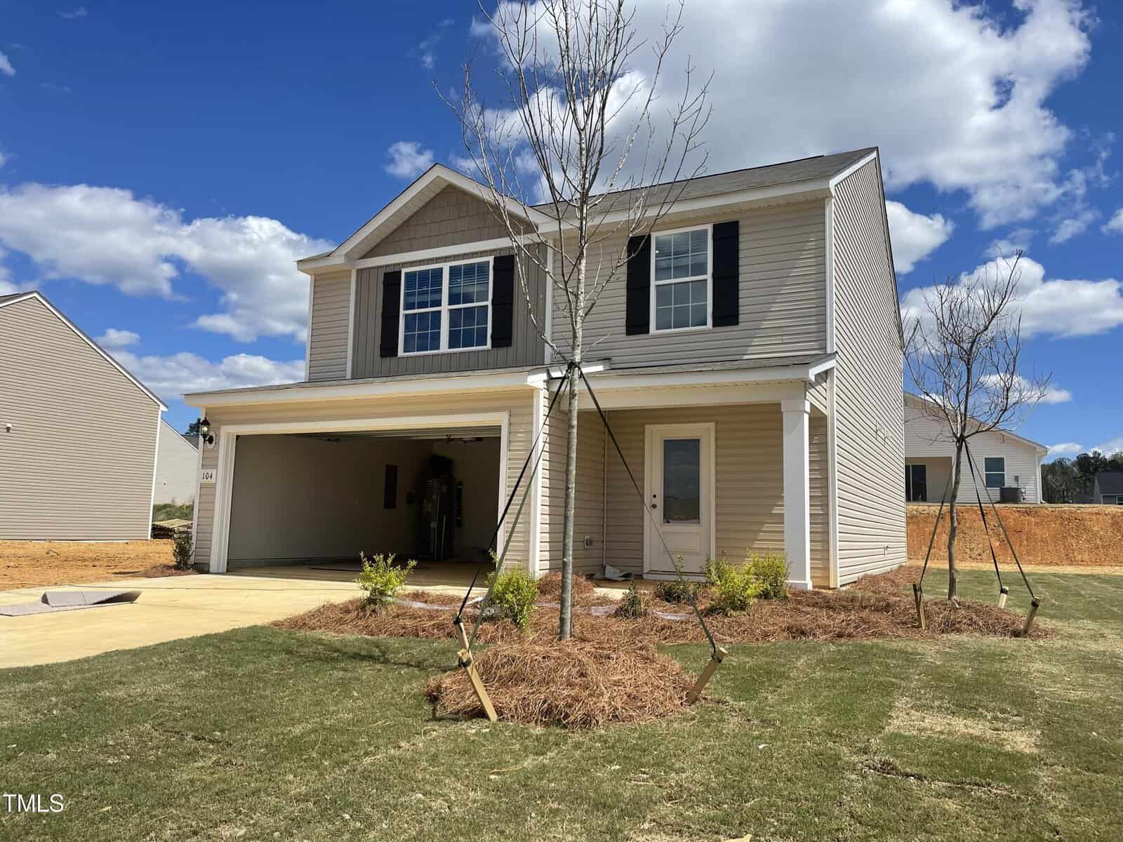 Nestled on 104 Anaheim Street, this two-story beige house in Oxford boasts a two-car garage and a quaint front yard. The open garage door reveals an empty space. Above, the sky is clear with a few fluffy clouds, while young trees and bushes adorn the landscape.