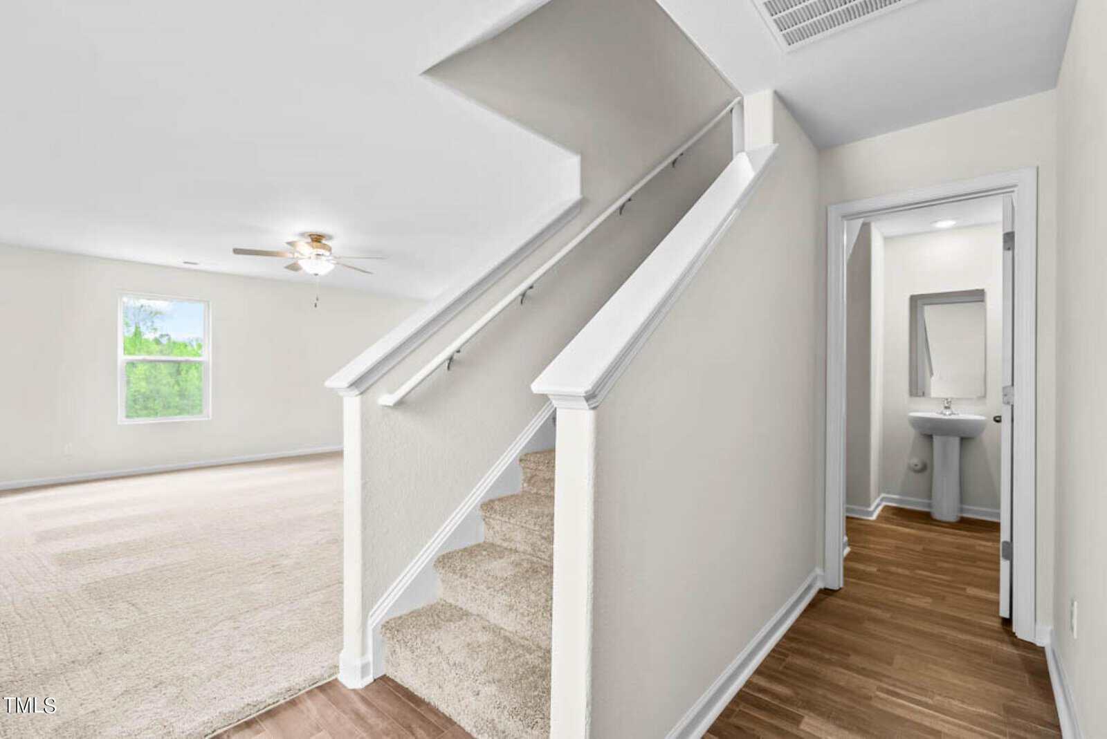 A bright, modern interior featuring carpeted stairs with a white railing leading to an open space. The room has beige carpeting, a window, a ceiling fan, and a small hallway with a wooden floor leading to a bathroom with a pedestal sink and mirror.