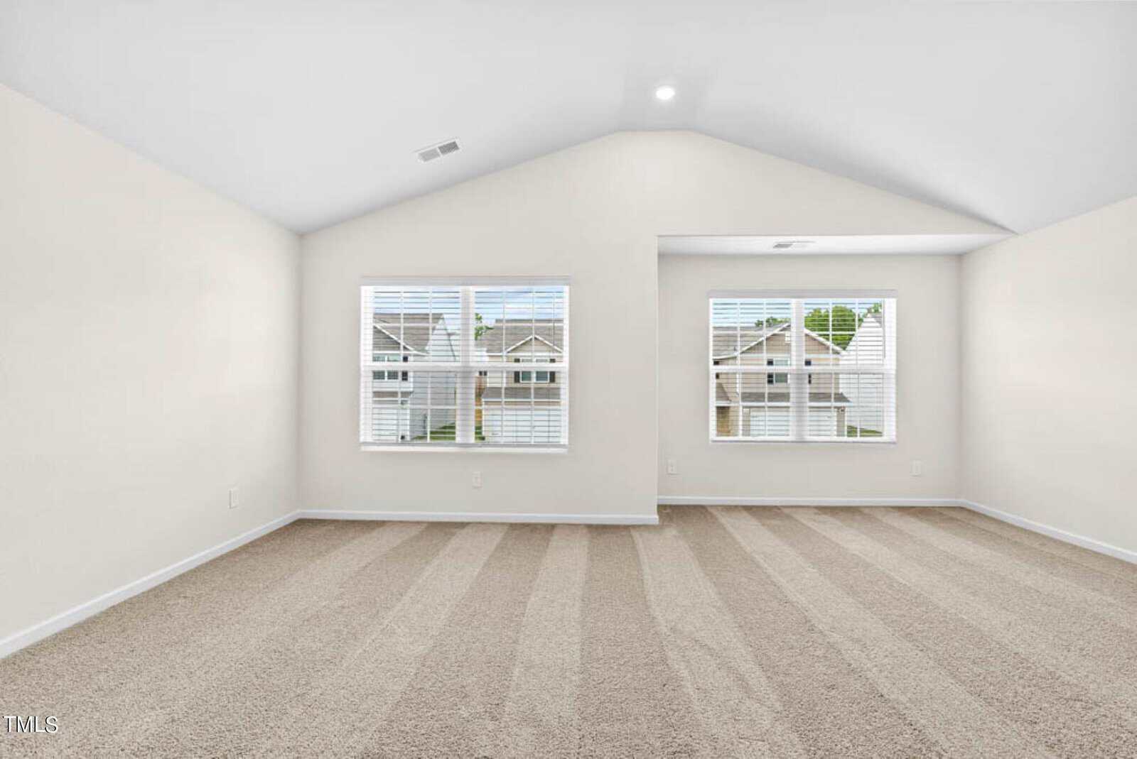 Spacious empty room with beige carpet, white walls, and a vaulted ceiling. Two large windows allow natural light to flood in, showcasing a view of neighboring houses.