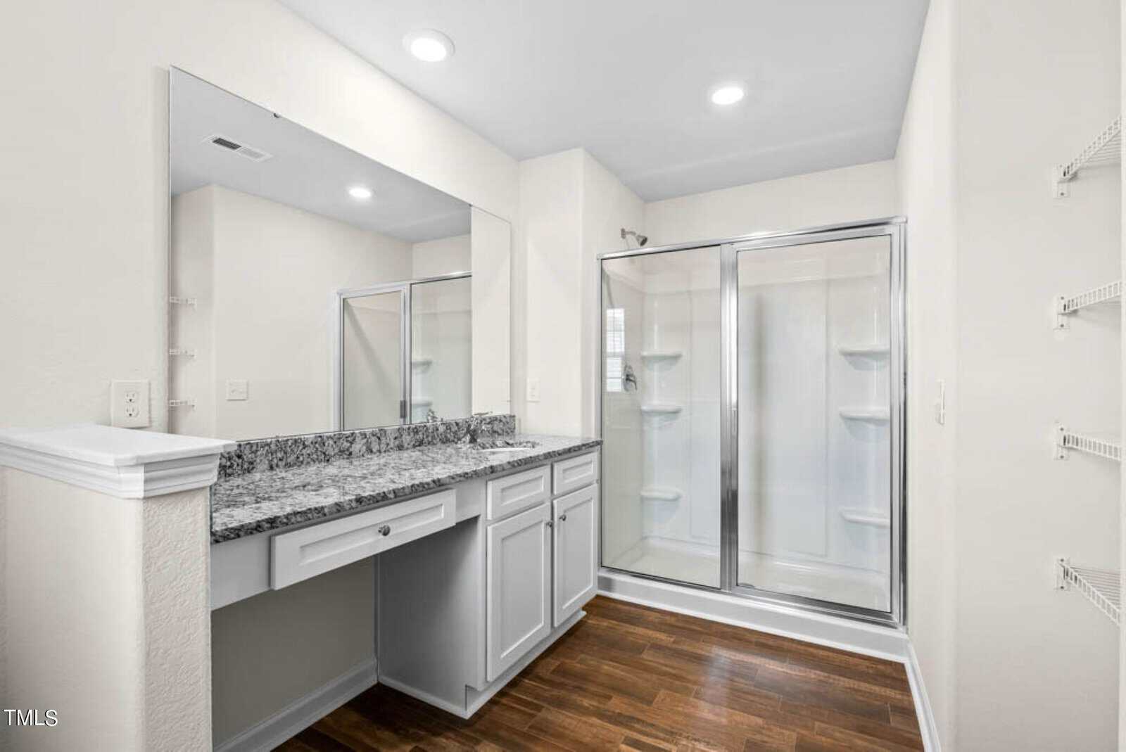 Modern bathroom with a large mirror above a granite countertop, white cabinets, and a glass-enclosed shower. The floor is wooden, and the walls are painted light beige. Shelving is visible to the right.
