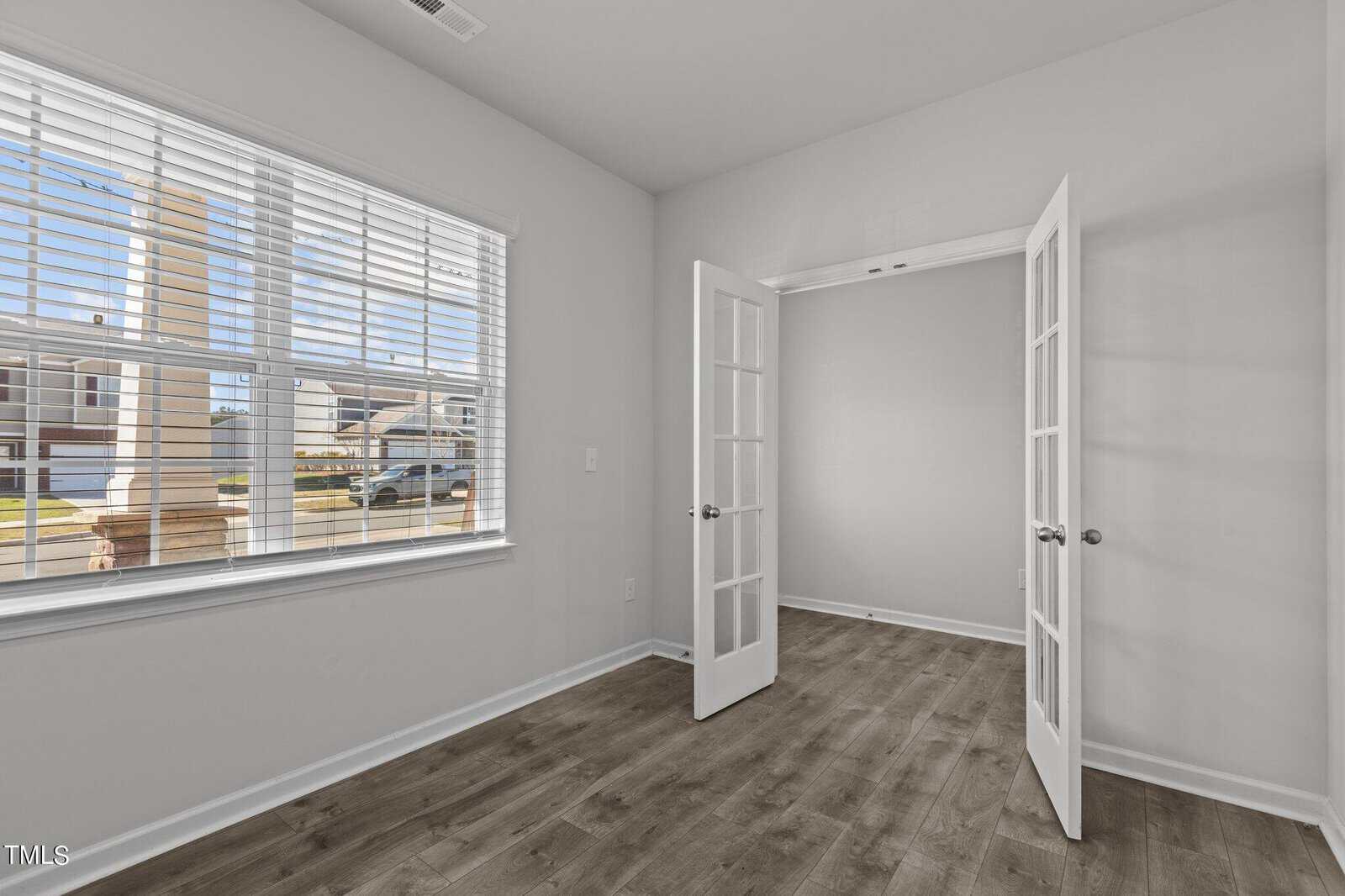 A bright, empty room with light gray walls and wood flooring. Large window with horizontal blinds on the left, allowing natural light in. Glass-paneled double doors open to another room.