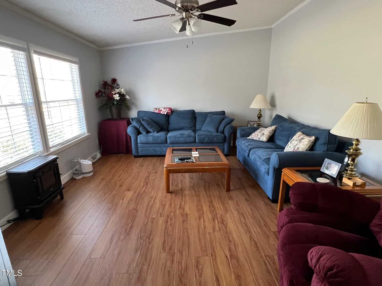 A living room featuring two blue sofas, a maroon armchair, a wooden coffee table, and a tv stand. the room has laminate flooring, a ceiling fan, and natural light from a window.