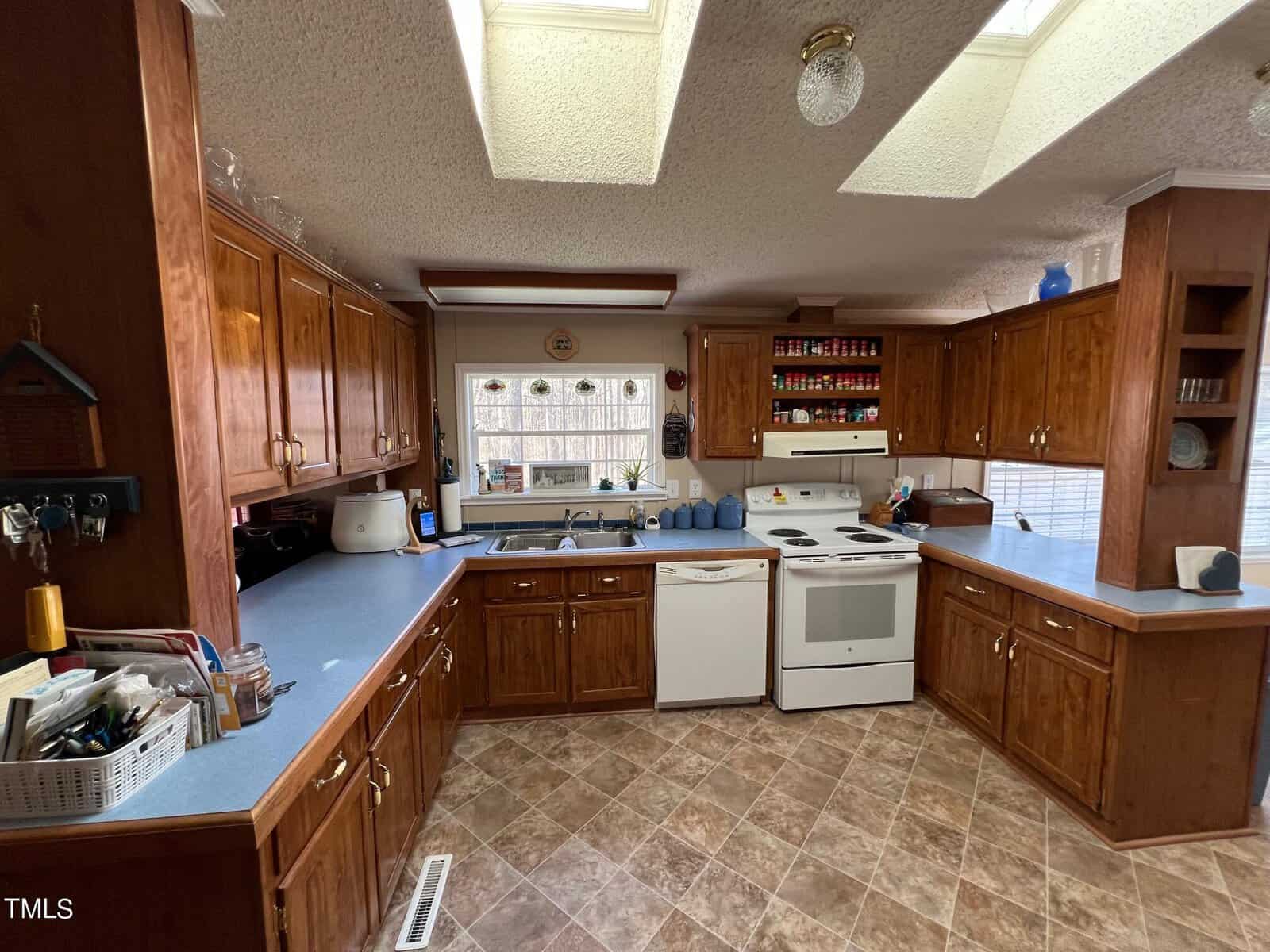 A spacious kitchen with wooden cabinets, tile flooring, and white appliances. features include a window overlooking a yard, a ceiling fan, and fluorescent lighting.