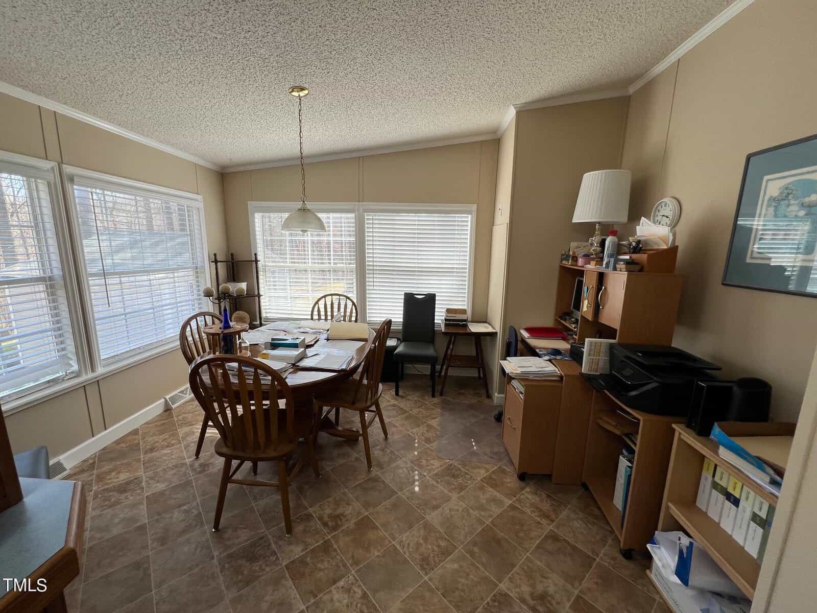 A spacious home office with beige walls, large windows with blinds, a dining table with chairs, a desk with a computer, shelves filled with books, and various office supplies scattered around.