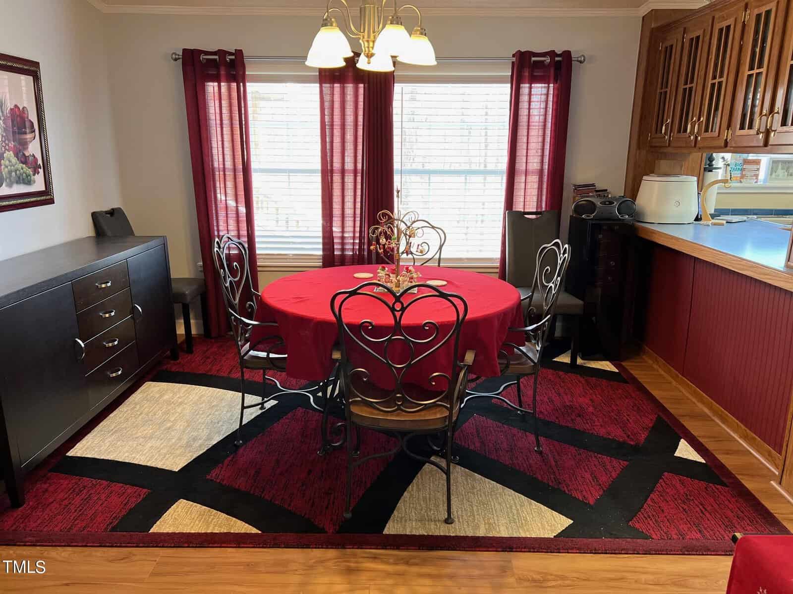 A cozy dining room with a round red tablecloth on a wooden table, surrounded by four chairs with heart-shaped backs. features include a patterned rug, burgundy curtains, and a view into a kitchen with wooden cabinetry.