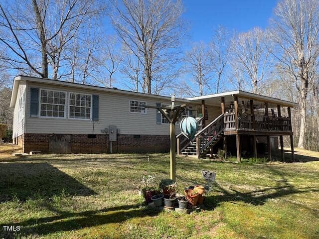 A single-story house with a raised wooden deck on the right, surrounded by bare trees and a grassy lawn. there's a basketball hoop near the deck under a clear blue sky.