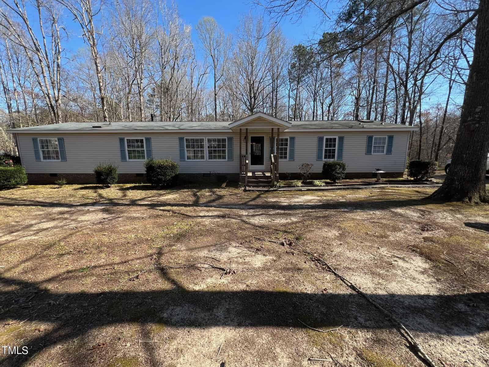 A one-story modular home located at 321 Springwood Drive, Henderson NC, with a light beige exterior and white trim, featuring a central entrance and symmetrical windows. The home is surrounded by bare