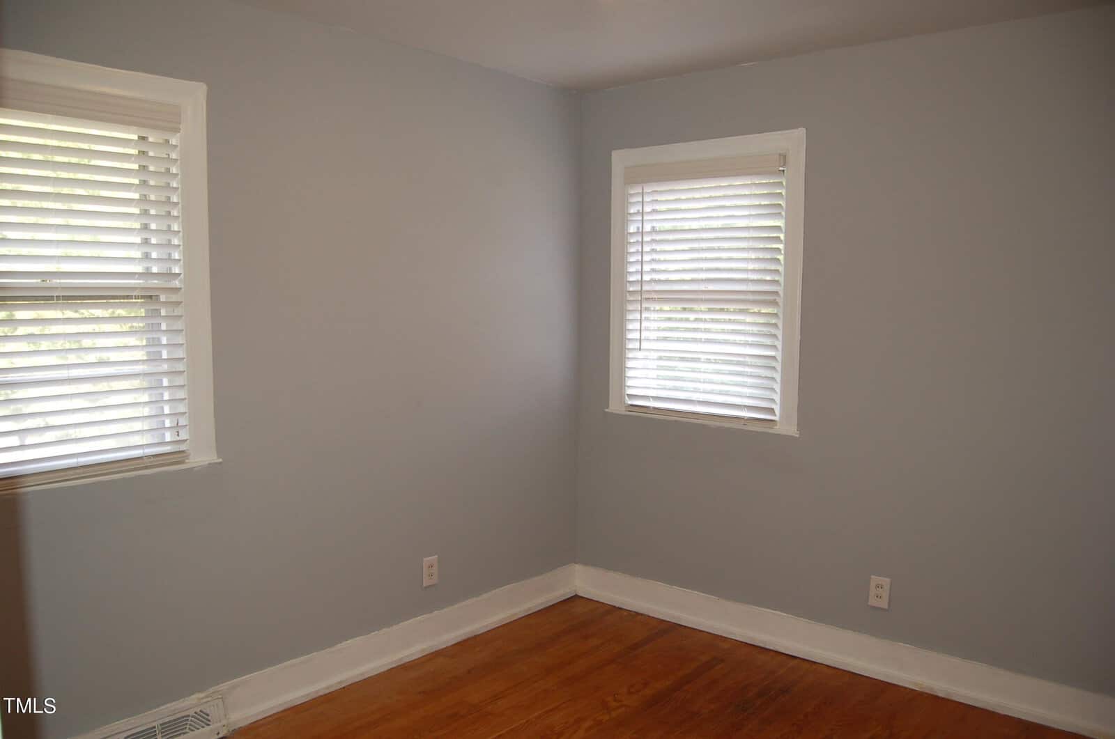 An empty room with light gray walls, two windows with white blinds, and a wooden floor. There's a baseboard and a vent on one wall.