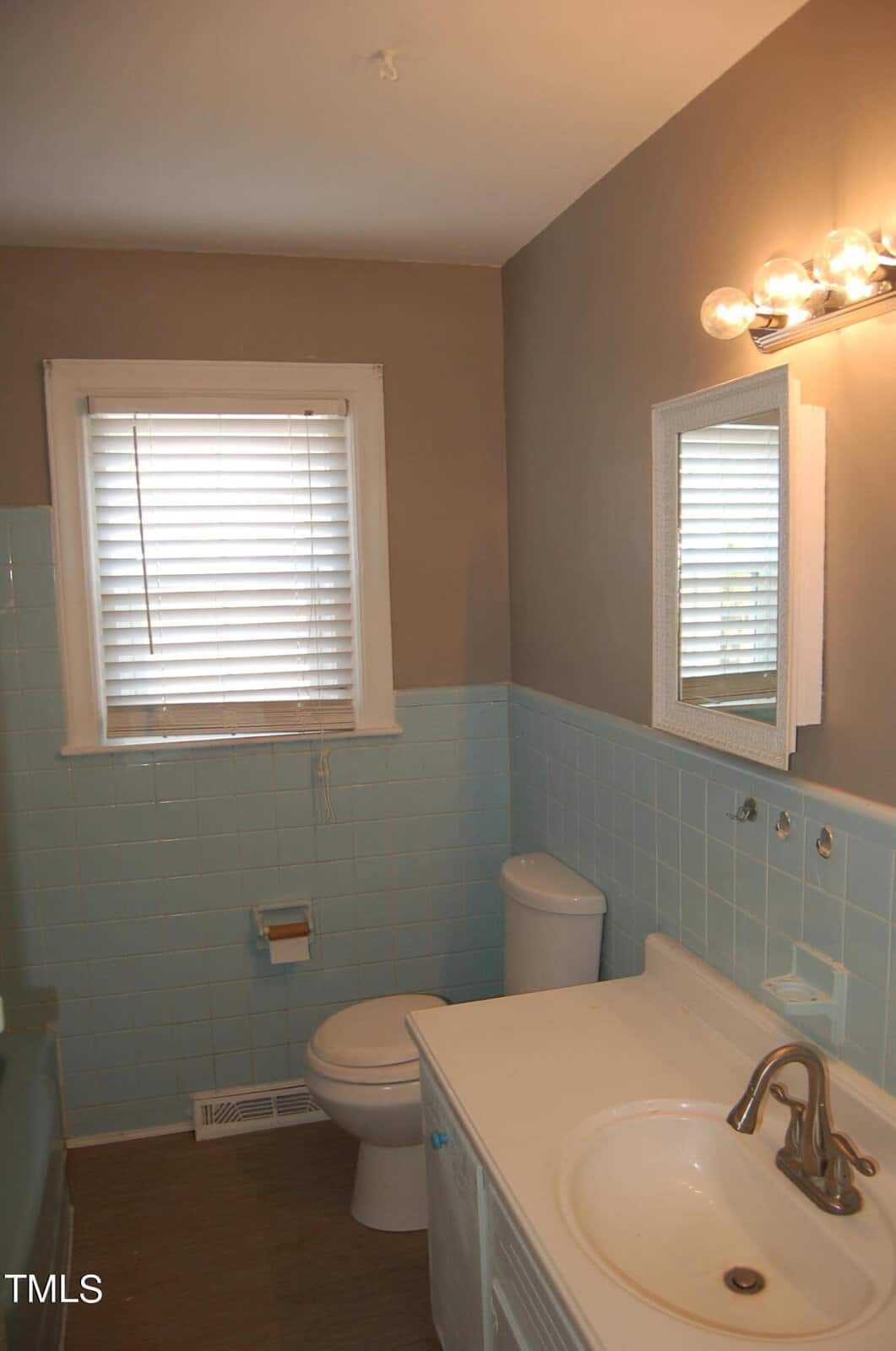A small bathroom with light blue tile walls and a beige upper wall. It features a white toilet, a white sink with a silver faucet, a rectangular mirror, and a row of round light bulbs above. A window with blinds is on the left.