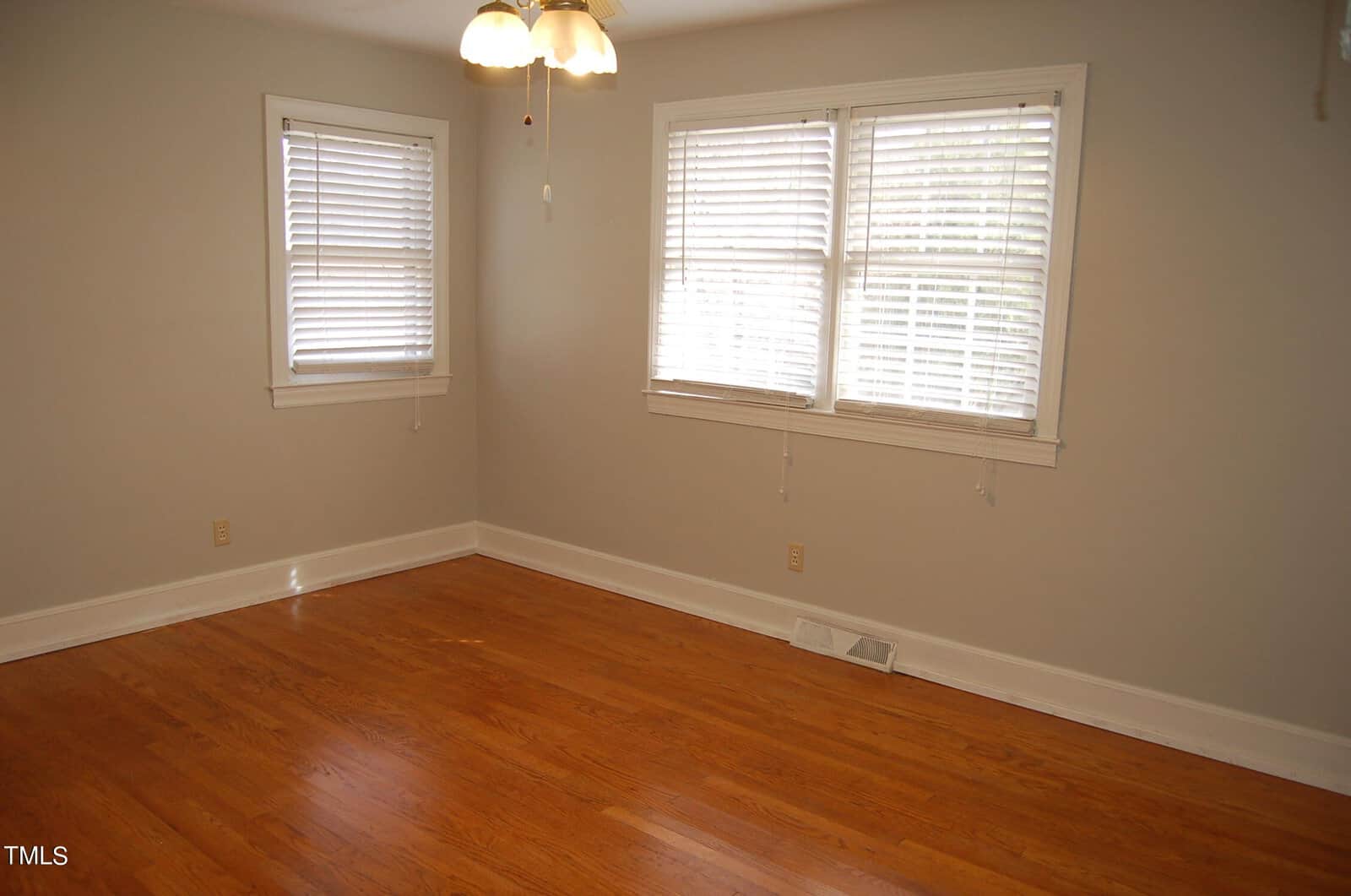 An empty room with light gray walls and hardwood floors. Two windows with white blinds allow natural light to enter. A ceiling fan with a light fixture is in the center of the ceiling.