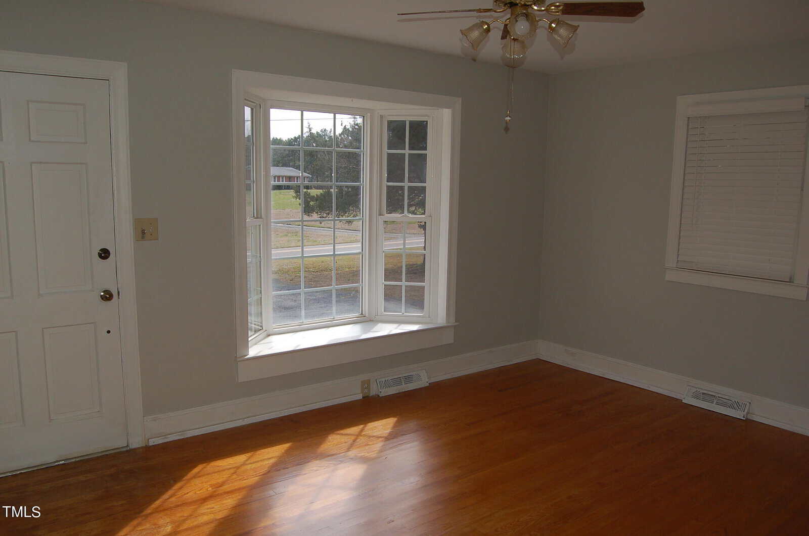 An empty room with light gray walls and hardwood floors. A large bay window with multiple panes lets in daylight. A white door is on the left, and a ceiling fan with lights is above.