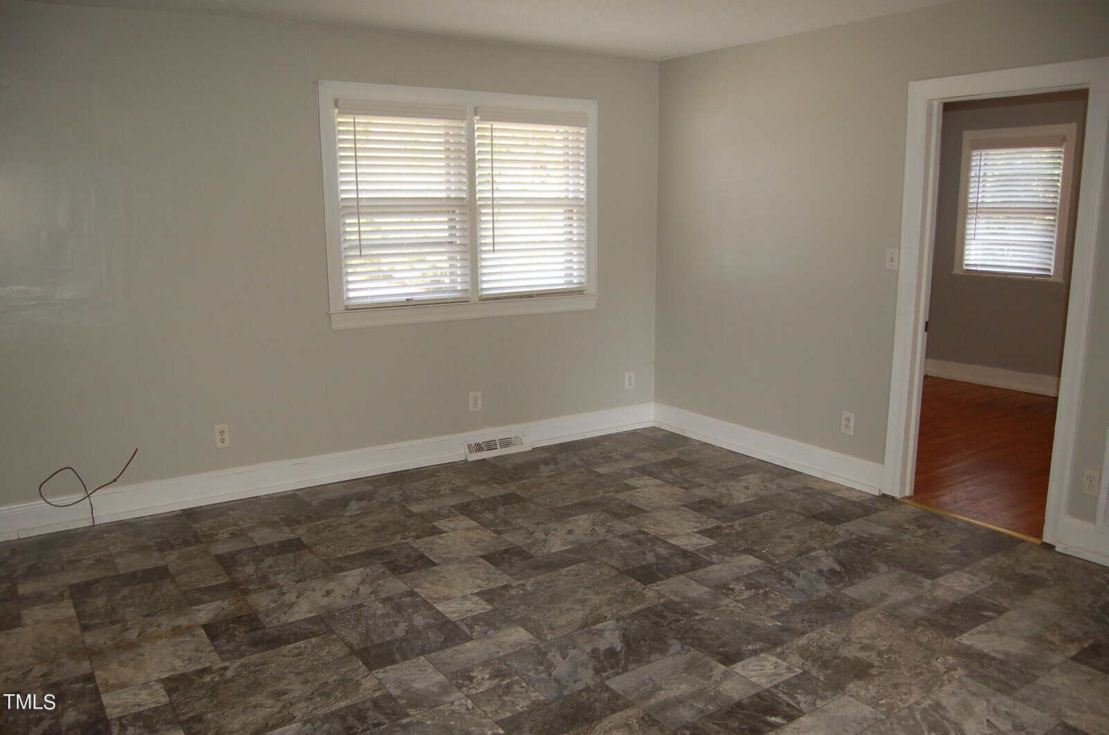 A room with light gray walls and a window with blinds. The floor is covered in gray and beige tile. An open doorway leads to another room with wooden flooring.