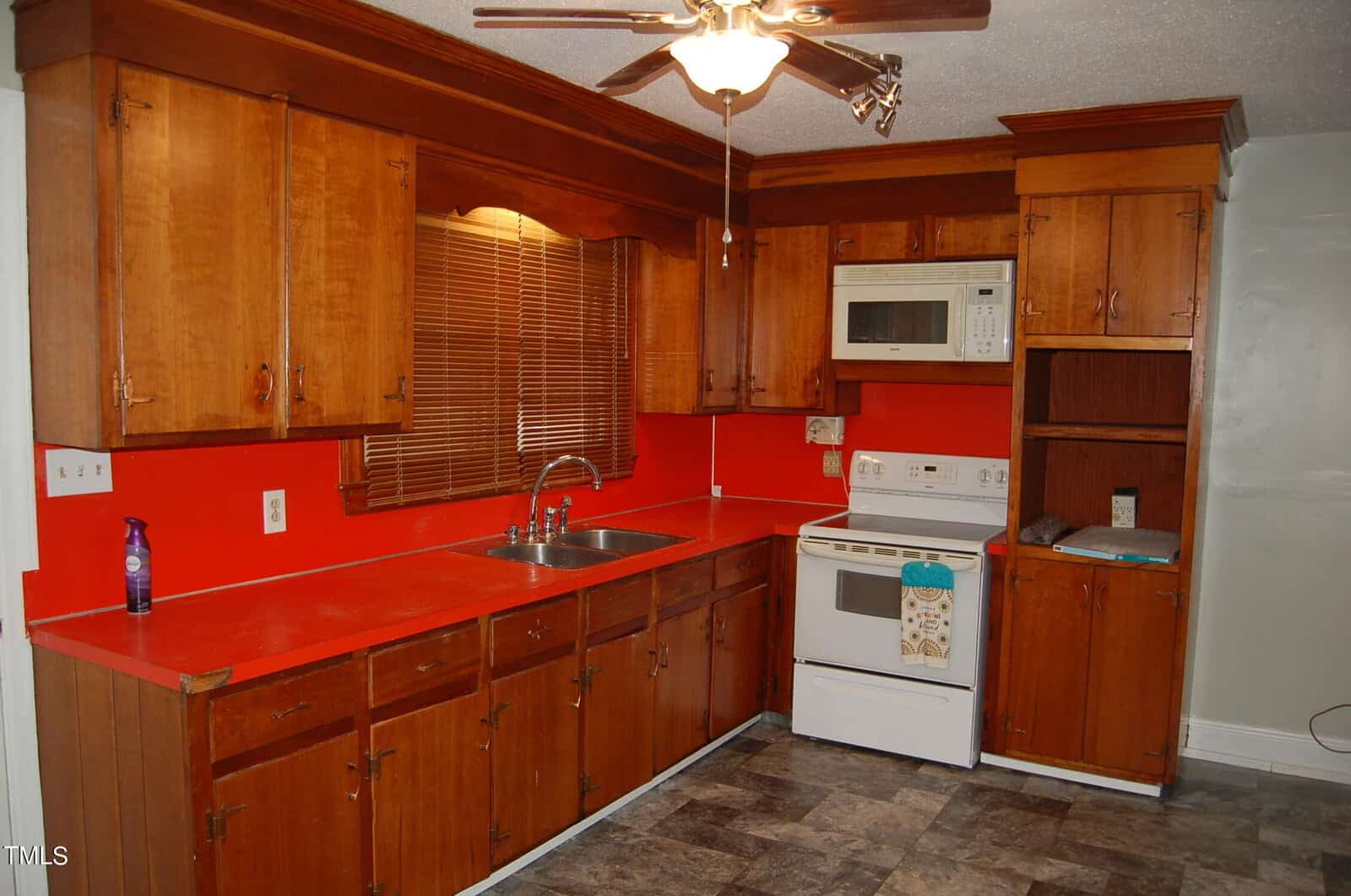 A kitchen with wooden cabinets and a red countertop. A ceiling fan with lights is above. The kitchen features a white stove with an overhead microwave, a stainless steel sink, and a red backsplash. A window with wooden blinds is present.
