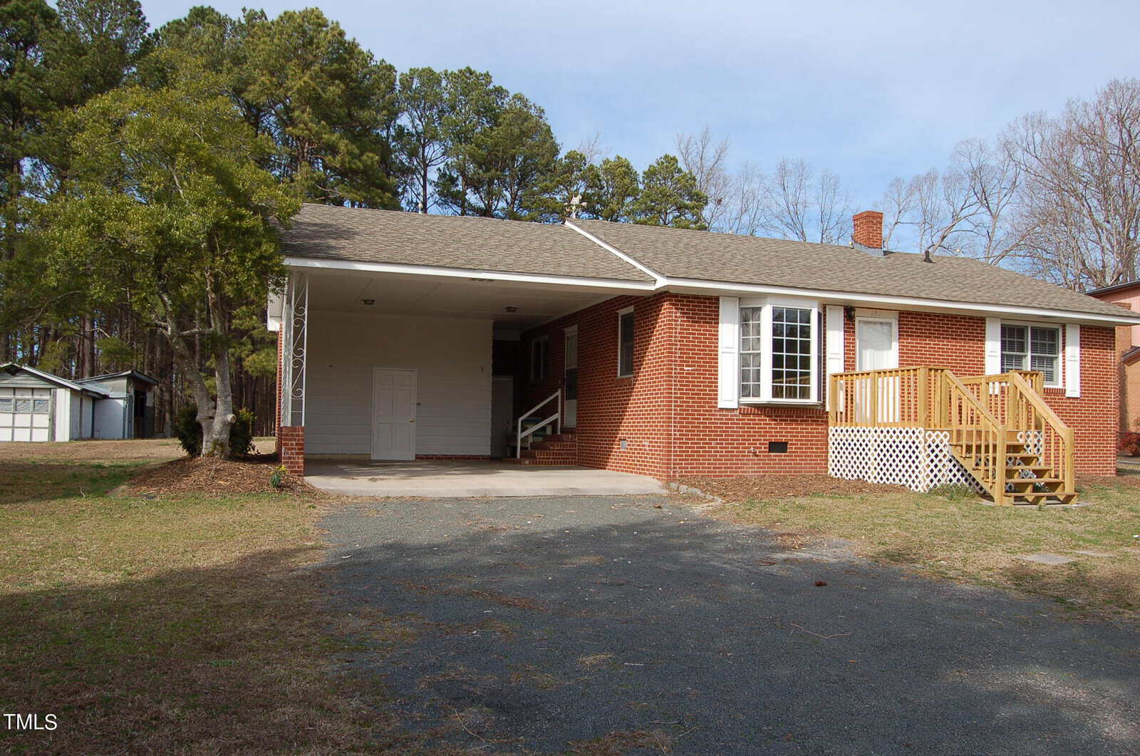 A brick single-story house with a carport on the left. The home features a covered porch with a few steps leading up to the front door. It is surrounded by a grassy yard and has trees in the background under a clear sky.