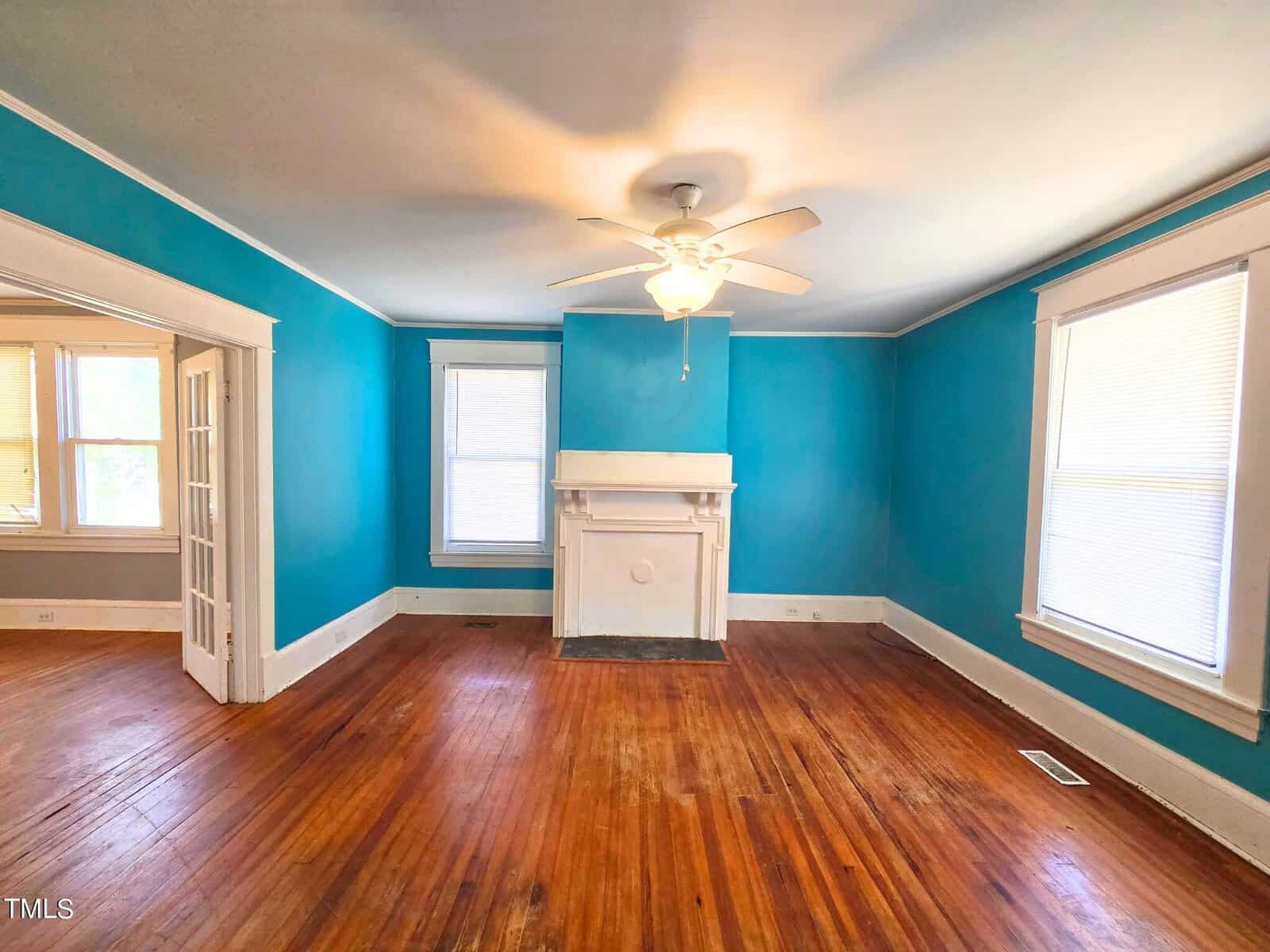 A bright living room with blue walls and a wooden floor. It features a ceiling fan light and a white fireplace centered between two windows. The room has French doors leading to another area with additional windows.