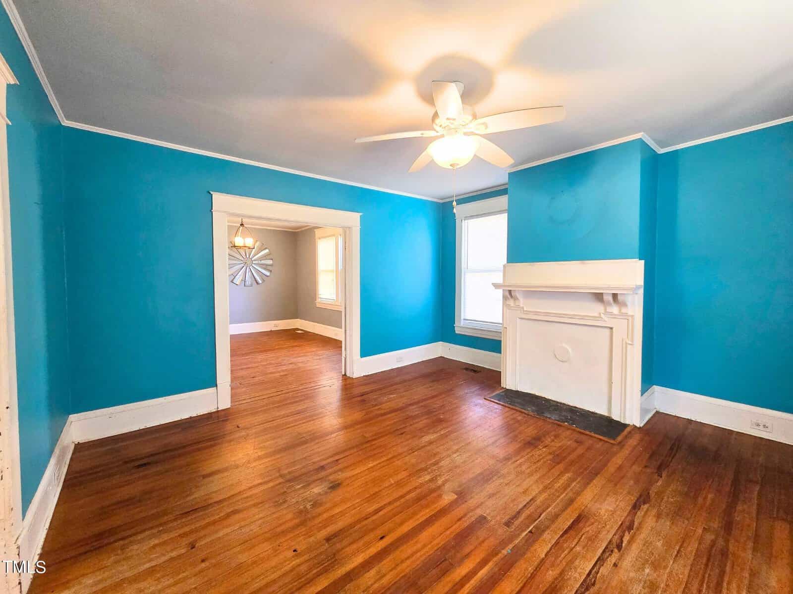 A bright living room with turquoise walls, a white ceiling fan, and a decorative white fireplace. The wooden floor adds warmth. A doorway leads to an adjacent room with gray walls and a decorative starburst on the far wall.