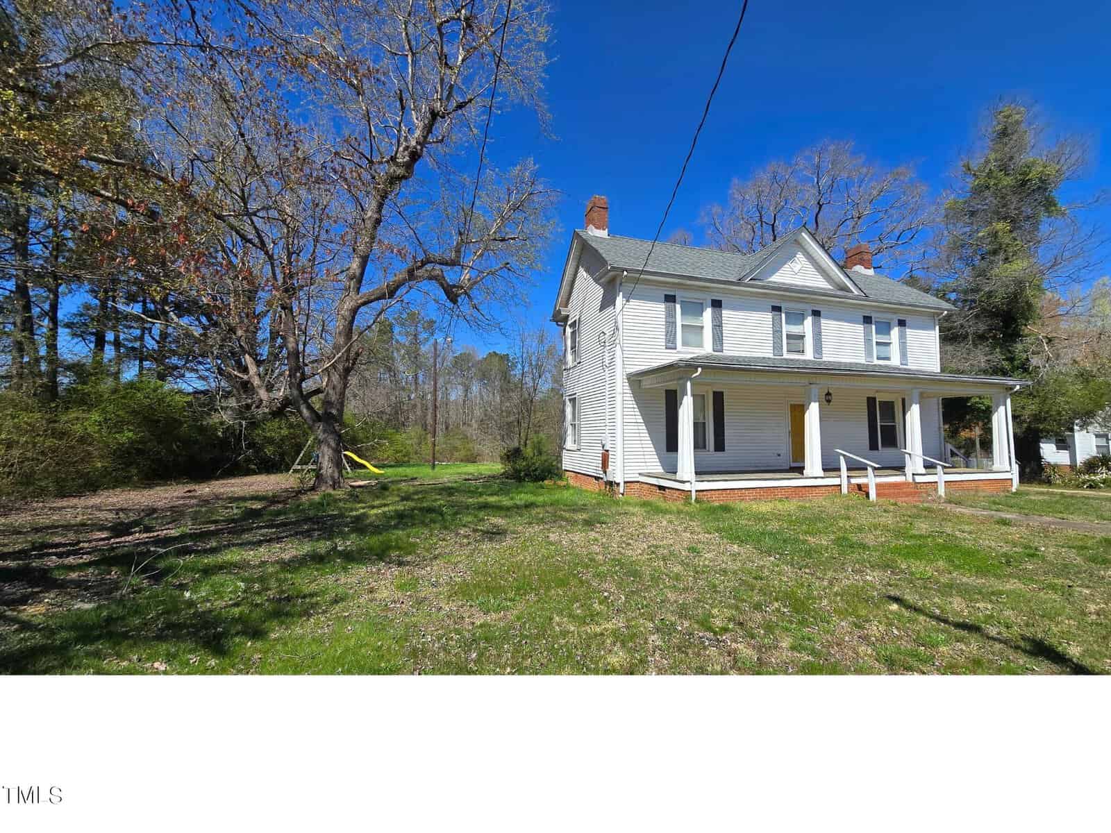 A two-story white house with a porch sits under a clear blue sky. The yard features a large, leafless tree on the left, with a yellow swing hanging from a branch. The background shows a mix of trees and open grassy areas.