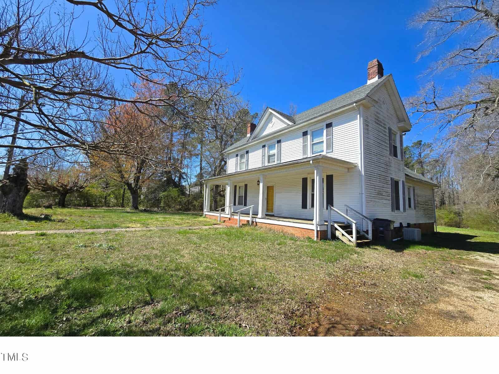A large, two-story white house with a front porch is surrounded by a grassy yard and bare trees. The sky is clear and blue, suggesting a sunny day.