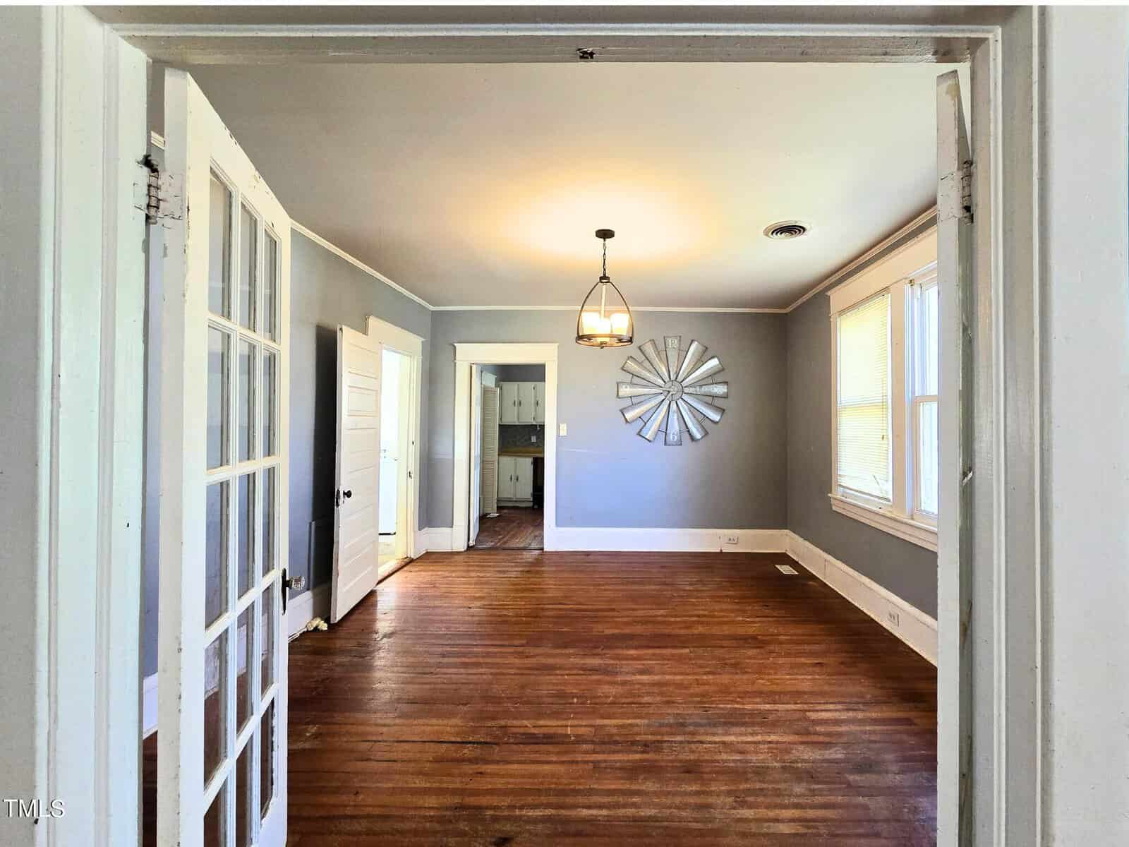 A room with wooden floors, gray walls, and a central light fixture. French doors open to the space, which features a metal wall decoration and leads to another room with white doors and trims. Large windows allow natural light in.