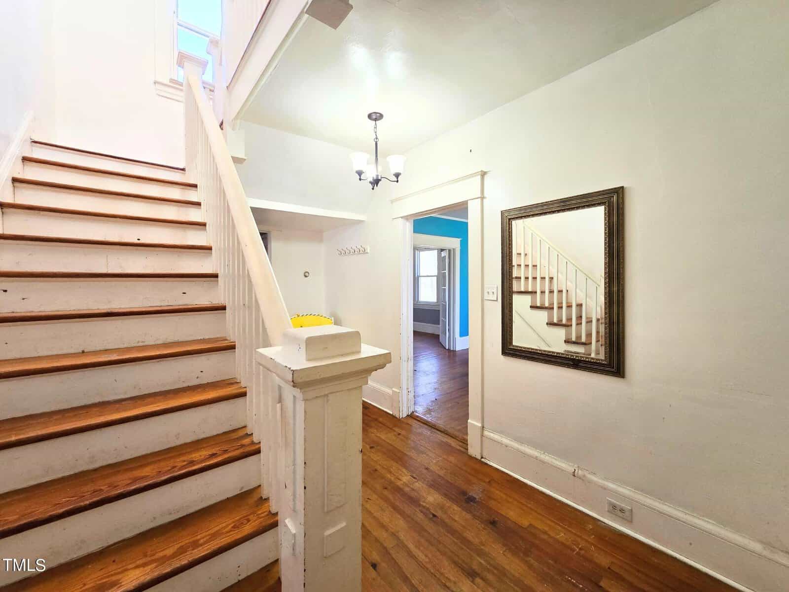 A bright hallway with wooden stairs to the left and a mirror on the right reflecting the staircase. A chandelier hangs from the ceiling, and there's a glimpse of a blue-walled room in the background. Natural light streams from a window above.