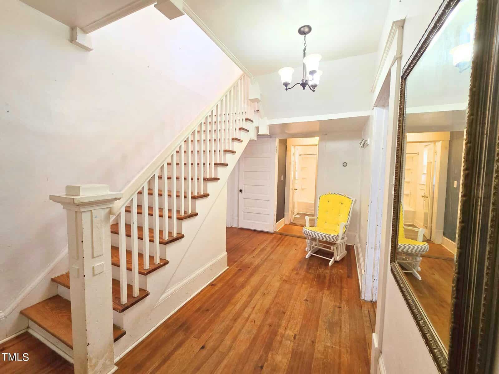 A hallway with wooden floors and a staircase on the left. A yellow chair is placed against the wall, and a chandelier hangs from the ceiling. There is a large mirror on the right wall reflecting the space.