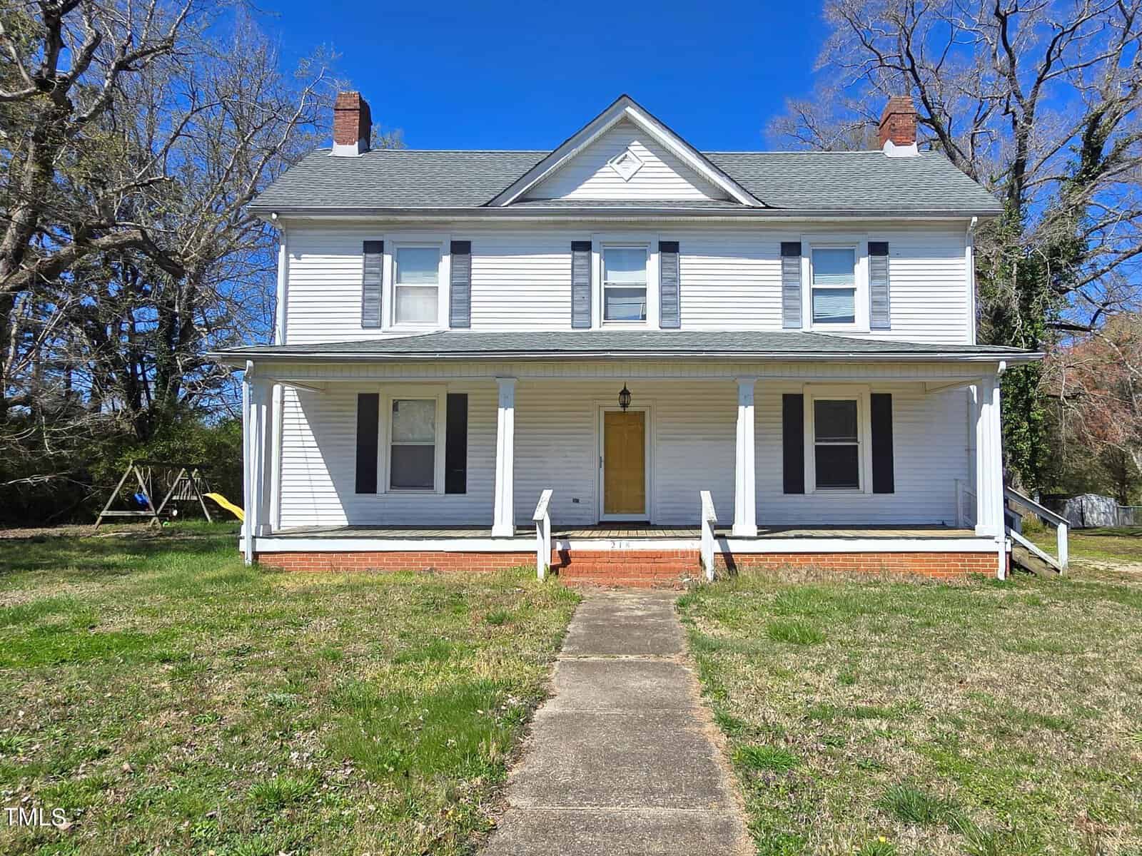 A two-story, white, wooden house with a covered porch, black shutters, and a peaked roof. The property has a path leading to the entrance, with grass and trees surrounding it under a clear blue sky.