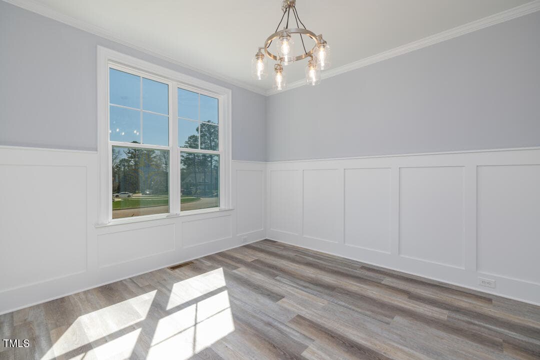 Empty room with gray walls, white wainscoting, and light herringbone-patterned floor. a modern chandelier hangs from the ceiling and sunlight streams through the large window.
