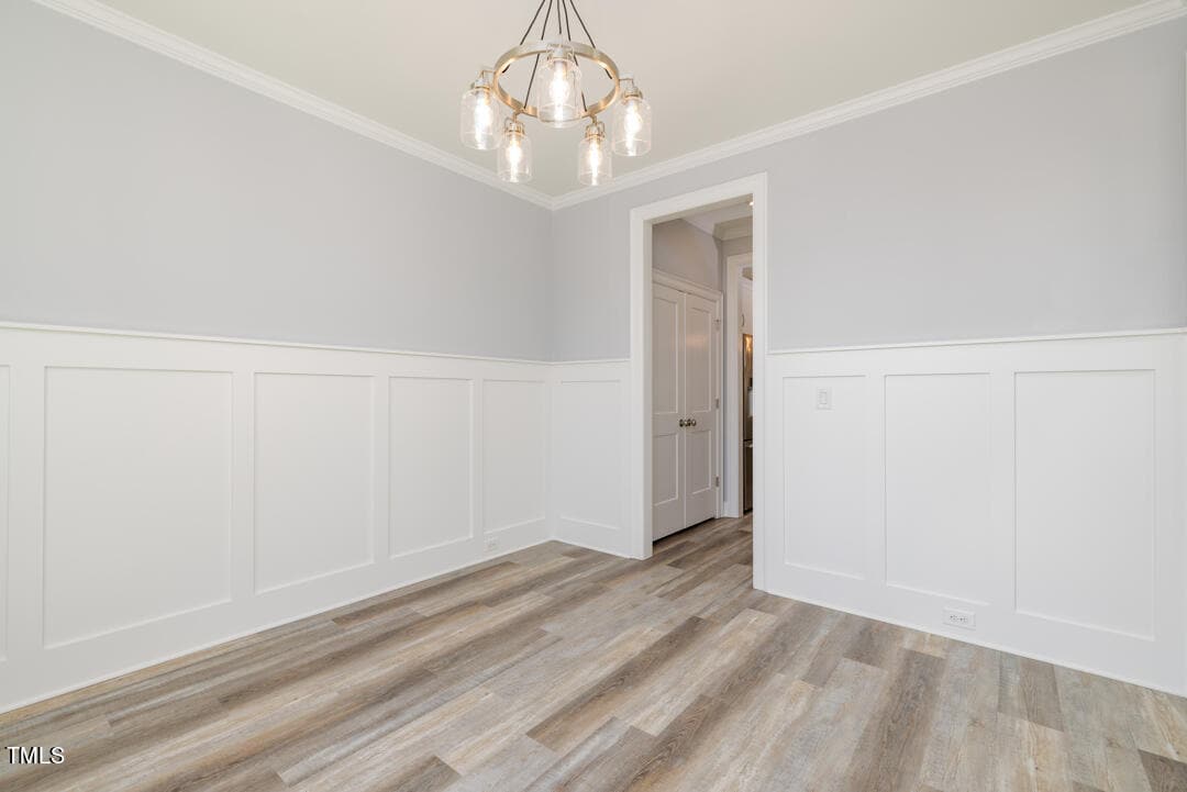 An empty room featuring gray walls with white wainscoting, light wooden flooring, and a chandelier hanging from the ceiling. an open door is visible in the background.