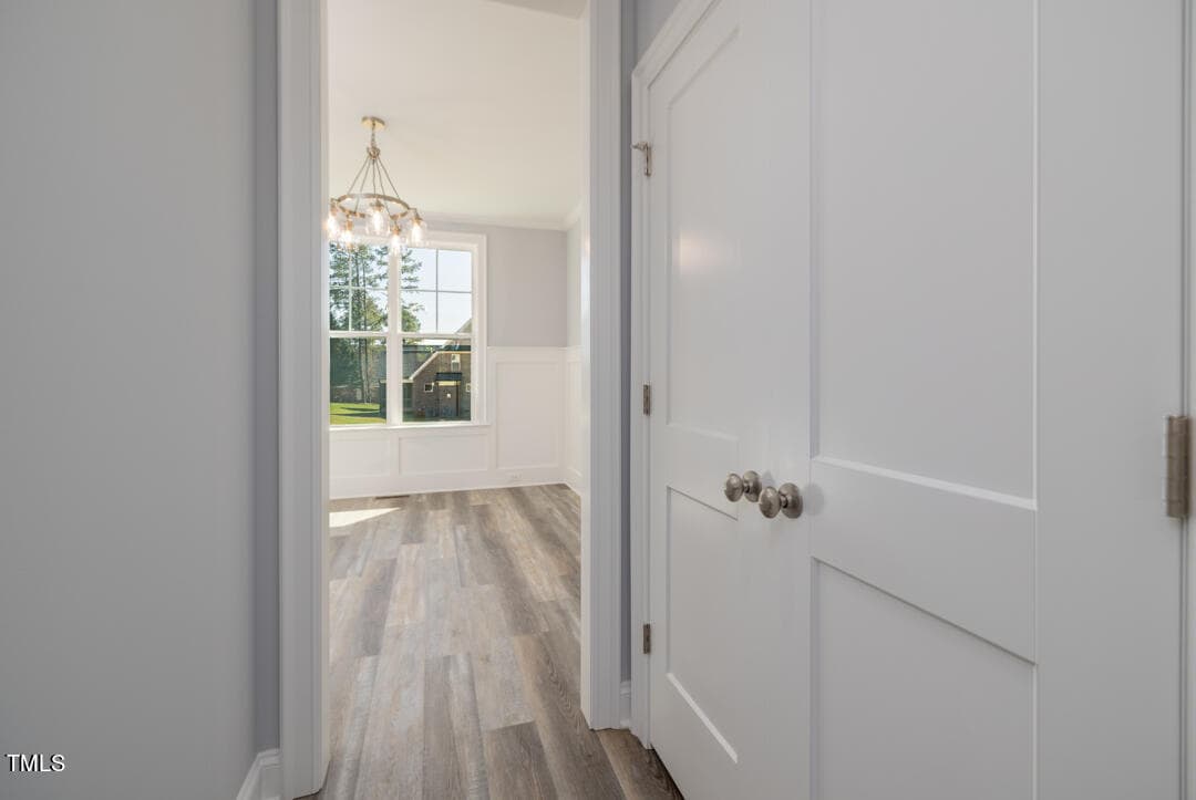 A bright, empty hallway in a house featuring white walls, wooden flooring, and a chandelier, with a view of windows at the end.