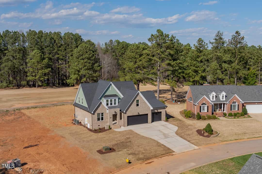 Aerial view of two detached houses with gabled roofs, one appears newly constructed on an unfinished landscape, next to a road with sparse traffic.