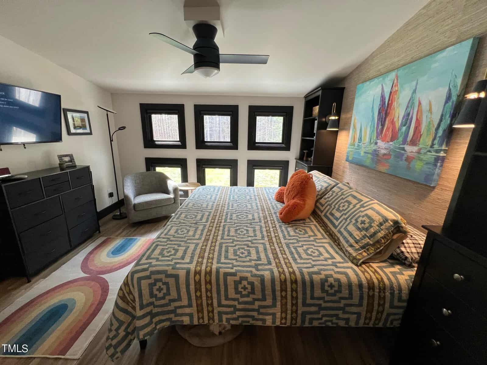 A cozy bedroom featuring a large bed with a geometric-patterned comforter, a vibrant painting of sailboats above the bed, dark furniture, and multiple windows.