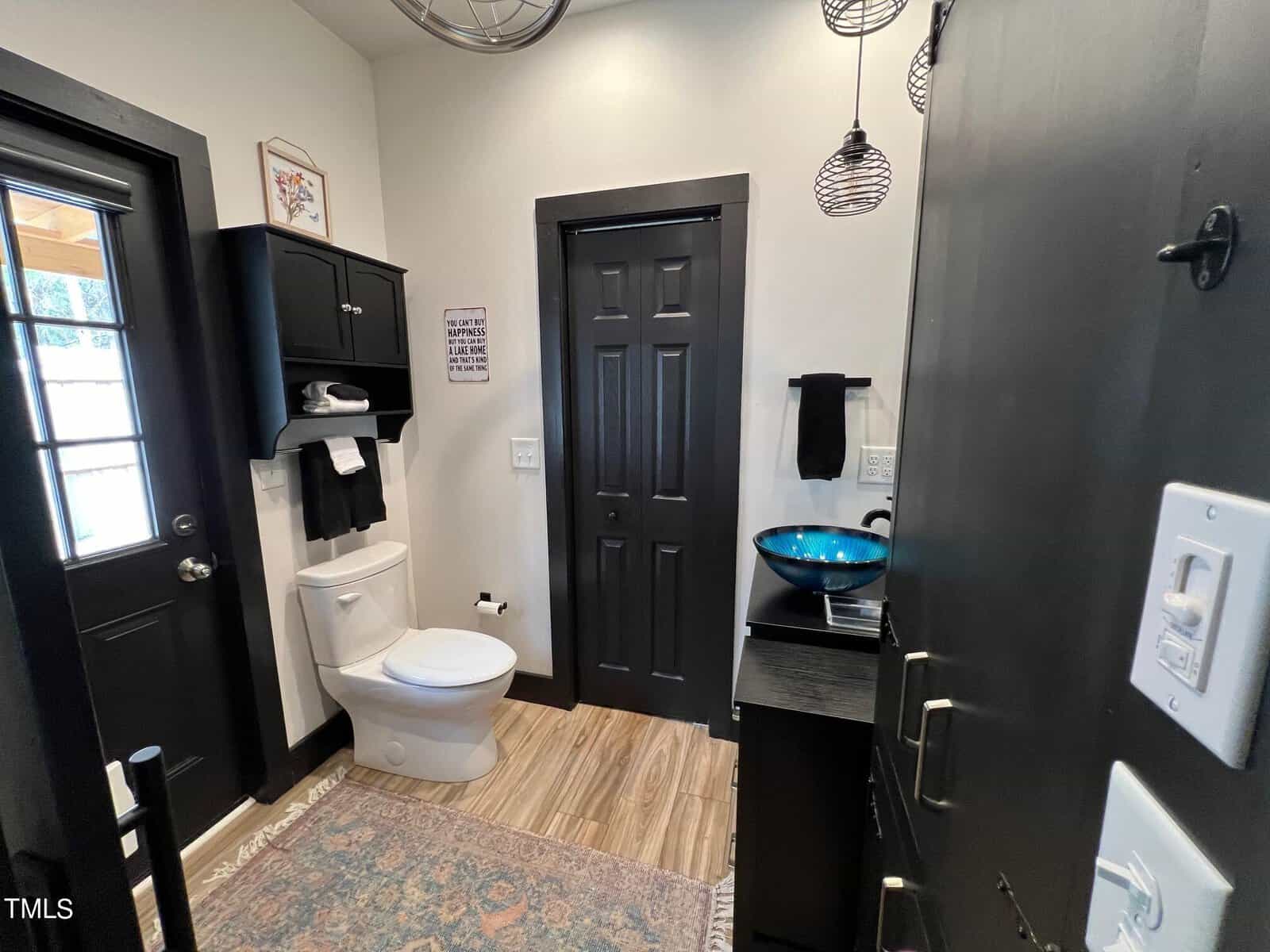 A stylish bathroom with black walls and a decorative blue bowl sink. features include a black door, white toilet, patterned rug, wall art, and hanging pendant lights.