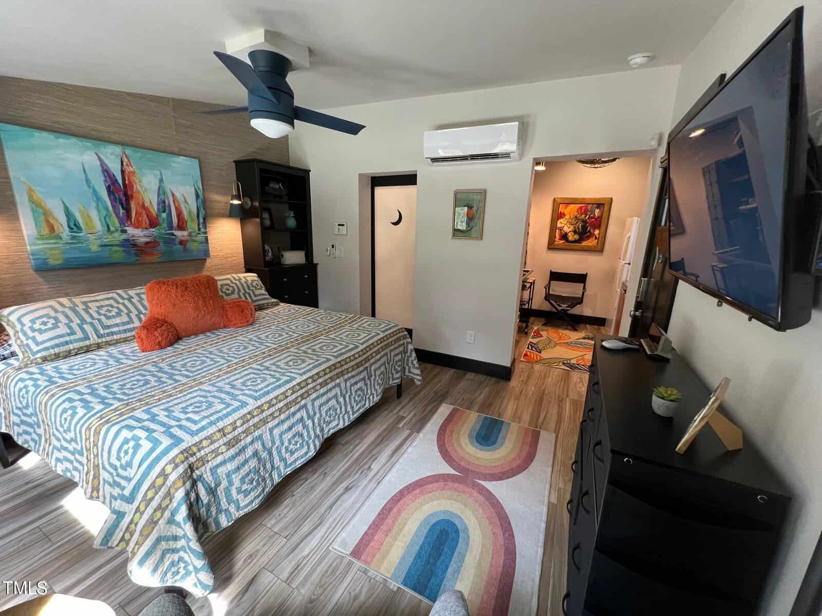 A modern bedroom with vibrant decor. there's a bed with a blue and white patterned cover, colorful paintings, a ceiling fan, dark furniture, and a mounted tv.