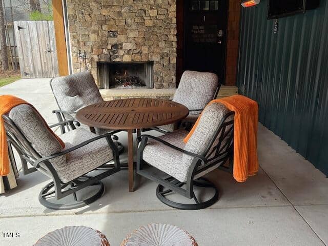 Outdoor patio setting featuring four cozy rocking chairs with gray cushions around a wooden table, orange throws on two chairs, and a lit fireplace in the background.