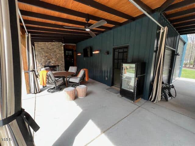 A cozy patio with a ceiling fan, furnished with two wicker chairs, a wooden table, and a stone fireplace. a glass door refrigerator stands on the right. sunlight illuminates the space.