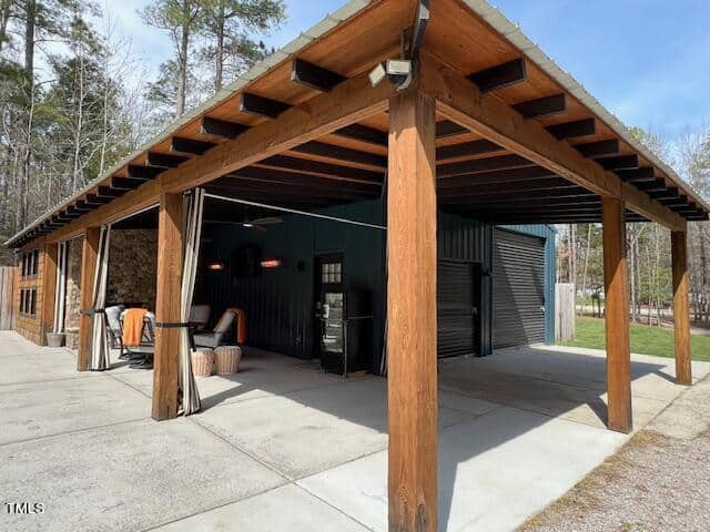 An outdoor shelter with a wooden frame and metal roof houses various tools and seating areas in a wooded setting, indicating a functional blend of storage and relaxation space.
