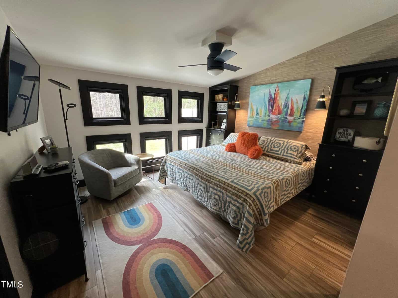 A modern bedroom with a bed covered in blue and white patterned bedding, colorful rug on the floor, dark brown trim windows, a fan overhead, and bright artwork above the bed. .