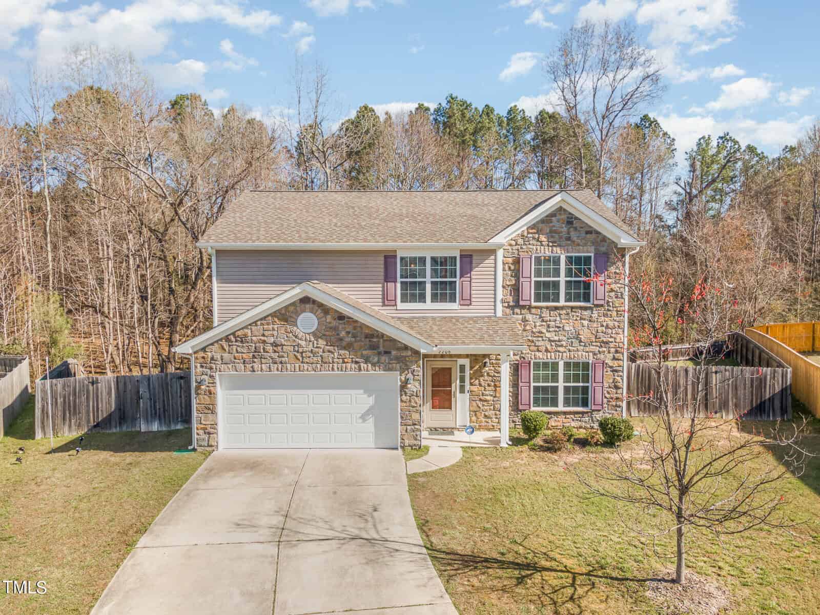 Located at 2206 Molton Court in Creedmoor, this two-story house features a stone facade, beige siding, and a two-car garage. A charming driveway leads through the front yard with its small tree. The home is enclosed by a wooden fence, all set against a backdrop of trees under a blue sky.